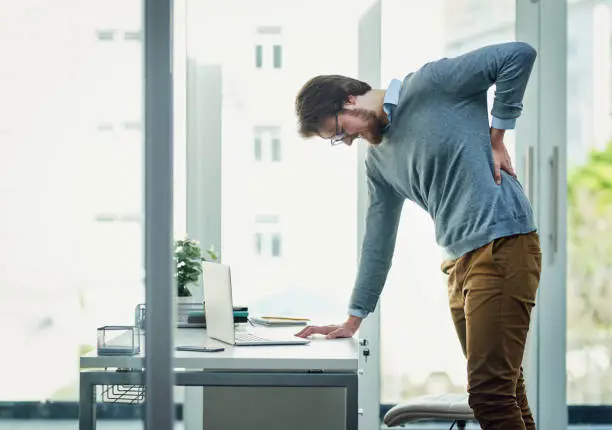Shot of a young businessman experiencing back pain while working at his desk in a modern office