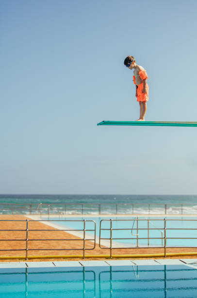 Boy learning to dive at swimming pool Boy with sleeve floats standing on high spring board and looking at swimming pool below. Boy learning to dive from diving platform at outdoor swimming pool. diving board stock pictures, royalty-free photos & images