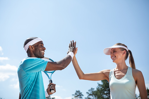 Low angle of positive couple playing tennis outdoor on warm sunny day. They are high-fiving while celebrating victory after tennis match. Athletes are standing beside while guy is holding racket
