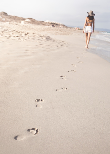 Distant Woman in shorts walking barefoot on white sandy beach in Formentera, Ibiza leaving footprints in sand. Focus is on the foreground footprints