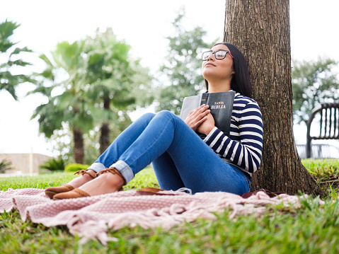 Pretty young woman sitting next to a tree and holding a bible close to her chest.