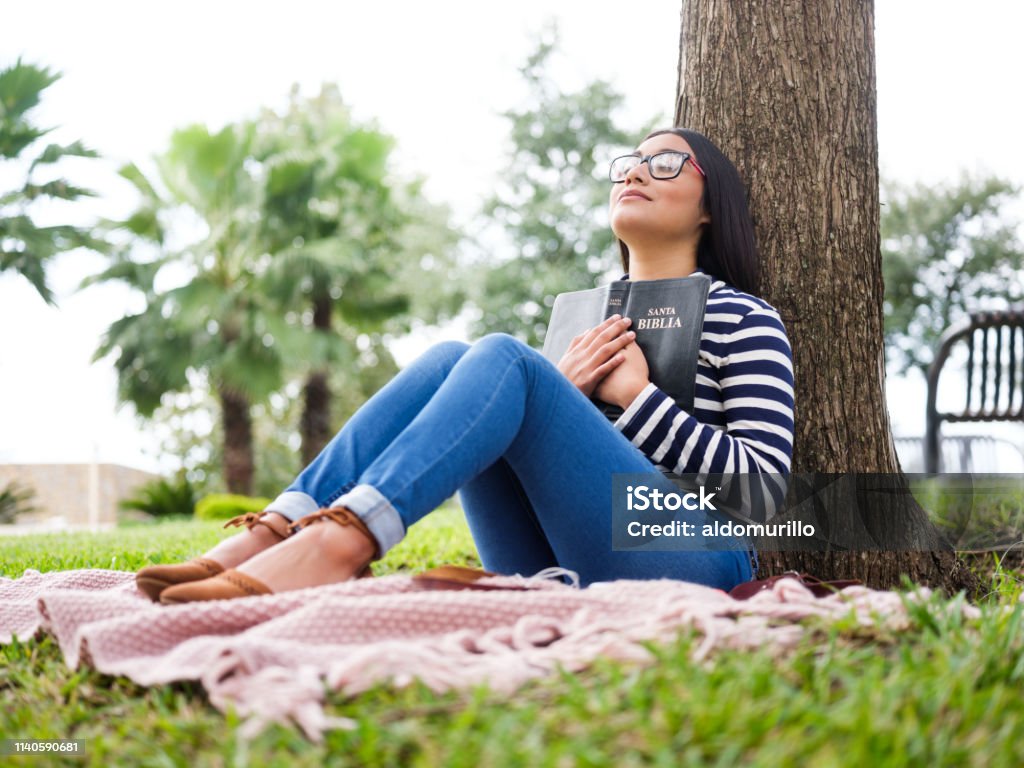 Pretty young woman sitting next to tree and holding Bible Pretty young woman sitting next to a tree and holding a bible close to her chest. Bible Stock Photo