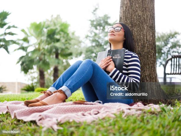 Pretty Young Woman Sitting Next To Tree And Holding Bible Stock Photo - Download Image Now