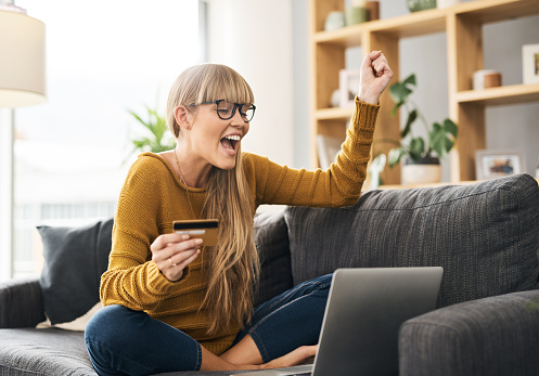 Shot of a young woman cheering while using a laptop and credit card on the sofa at home