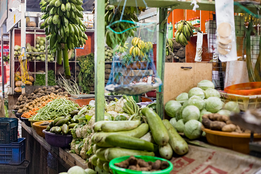 A vibrant display of vegetables, including striped eggplants, ridged gourds, and okra. A shopper, wearing a floral dress, carefully selects fresh okra, emphasizing the market's interactive atmosphere.