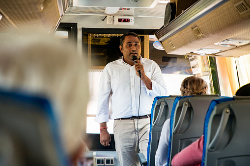 A front-view shot of a mid-adult indian tour guide coach driver talking to tourists using a microphone on a coach bus, the tourists are listening and they're ready to begin their journey.
