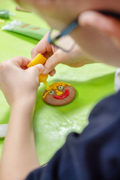 istock Decorating cinnamon cookies by a child. The boy's hands form a face of colored icing squeezed out of tubes. Unaddressed biscuits in the background. 1140629435