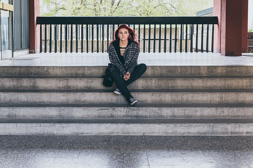 Young beautiful red hair girl sitting alone outdoors on the stairs of the building with hat and shirt feeling anxious and depressed after she became a homeless person