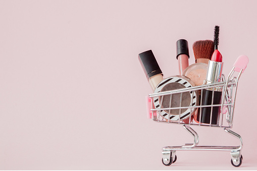 Concepto creativo con carrito de compras con maquillaje sobre fondo rosado. Perfume, esponja, cepillo, rímel, lápiz, Lima de uñas, sombra de ojos, brillo labial en la cesta, copiar espacio photo