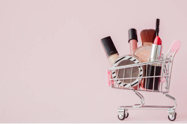 Creative concept with shopping trolley with makeup on a pink background. Perfume, sponge, brush, mascara, pencil, nail file, eye shadow, lip gloss in the basket, copy space