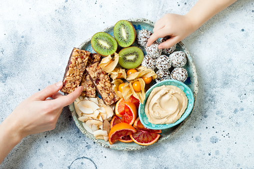 Mother giving healthy vegan dessert snacks to toddler child. Concept of healthy sweets for children. Protein granola bars, homemade raw energy balls, cashew butter, toasted coconut chips, fruits platter