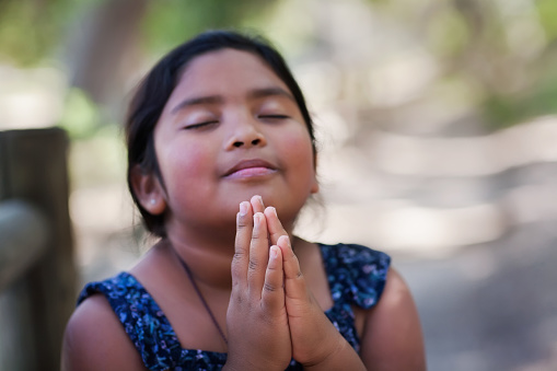 A native young girl with hands together in prayer, in an outdoor setting praying to God with a subtle smile.