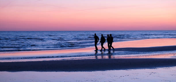 Four unrecognizable young men walking by the sea on a deserted sandy beach, they are close together talking there is a pink glow in the sky reflecting on the beach the sea is deep blue, Brighton, UK