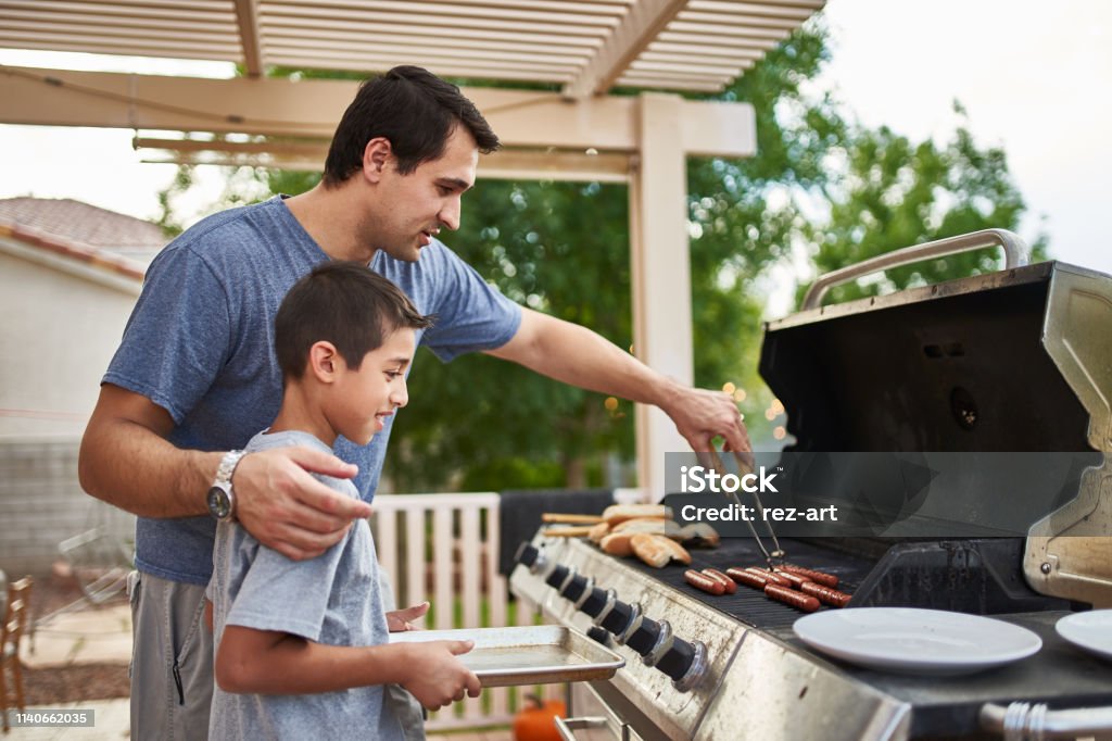 father teaching son how to grill hot dogs and bonding father teaching son how to grill hot dogs and bonding during the day Barbecue Grill Stock Photo