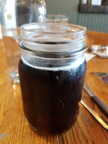 Close-up of glass jar of artisan root beer on tap, on a light wooden surface, April 3, 2019