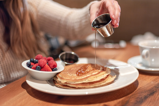 Closeup of young woman at cafe table pouring warm maple syrup on pancakes