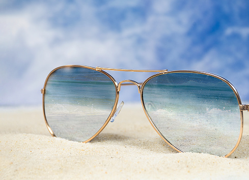 close up of aviator sunglasses in beach sand with ocean surf reflection