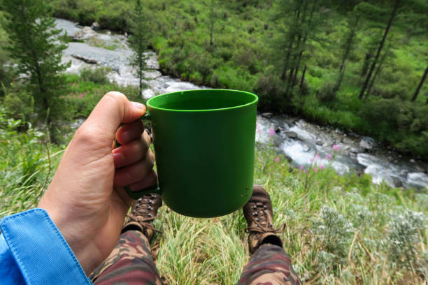 Tourist hand holding plastic mug. Camping image. Enjoying rest and mountain river and forest. Nomad life stock photo