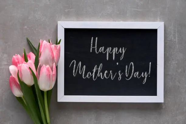 Greeting text "Happy Mother's day!" on blackboard with white tulips with pink stripes, flat lay on grey stone background