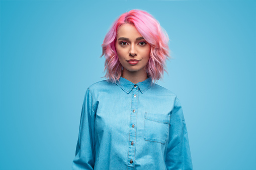 Beautiful young woman with trendy wavy pink hairstyle wearing blue shirt and looking at camera on blue background
