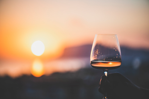 Man's hand holding glass of rose wine and with sea and beautiful sunset at background, close-up, horizontal composition. Summer evening relaxed mood concept
