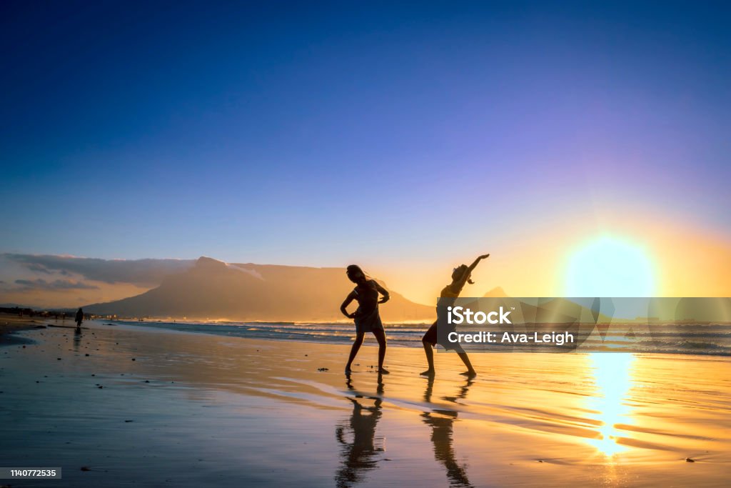 Two African women dancing on the beach at sunset, with Table Mountain and Cape Town in the background, Milnerton Beach, Cape Town, South Africa Dancing Stock Photo