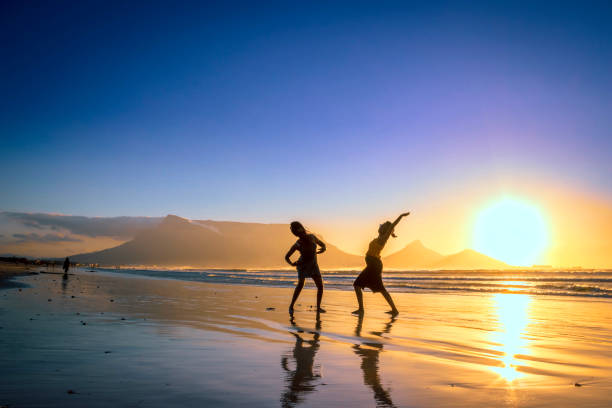 Two African women dancing on the beach at sunset, with Table Mountain and Cape Town in the background, Milnerton Beach, Cape Town, South Africa stock photo