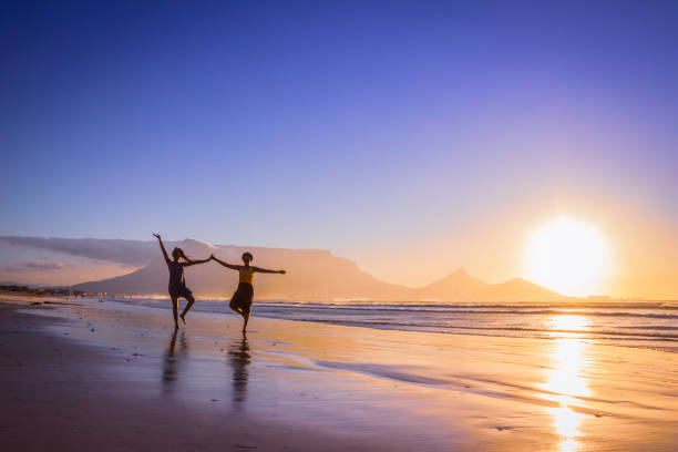 две африканские женщины танцуют на пляже на закате, с столовой горы и кейптауна в фоновом режиме, милнертон-бич, кейптаун, - milnerton стоковые фото и изображения