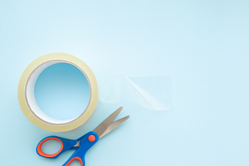 Transparent roll of scotch and scissors on light pastel blue background. Closeup. Empty place for text. Top view.