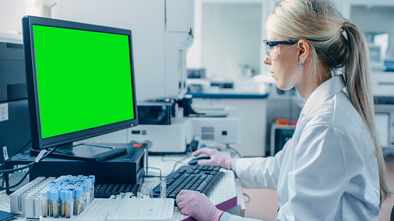 Female Research Scientist Sits at His Workplace in Laboratory, Uses Green Mock-up Screen Personal Computer. In the Background Genetics, Pharmaceutical Research Centre.