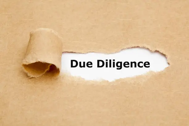 Text Due Diligence appearing behind torn paper. Concept representing the research done before entering into an agreement or contract as a part of the risk analysis process.