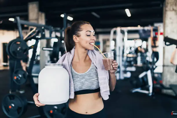Young attractive woman after successful workout posing and smiling in modern fitness gym while holding big white protein jar and glass of shake with drinking straw in other hand.