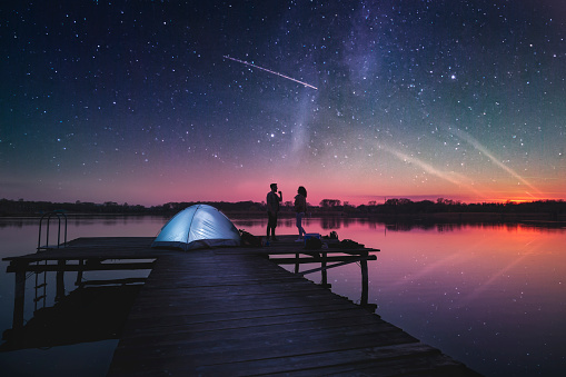 Combination of high ISO, noisy image of a starry sky and image of a couple camping on lake docks at dusk, standing next to a tent enjoying beautiful summer night