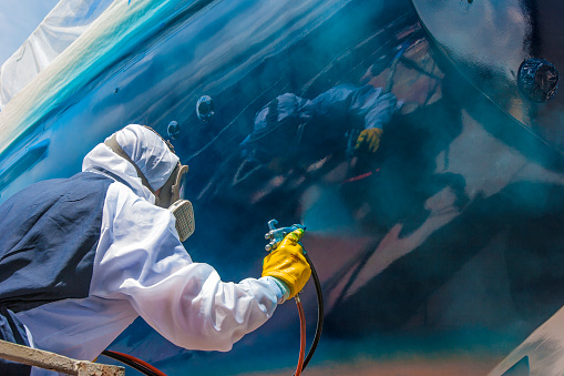 Whangarei, New Zealand, 11-30-2018, worker in full body protection suit and mask is spray painting the hull of a sailing yacht as final work of corrosion protection