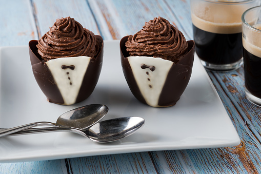 Chocolate mousse in delicate molded chocolate tuxedo cups on a white serving plate with two spoons. Two cups of dark espresso sit nearby  Plate is on a light blue wood table.