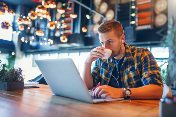 Photo of Young Caucasian blogger with earphones in ears and in plaid shirt drinking coffee and using laptop while sitting in coffee shop.