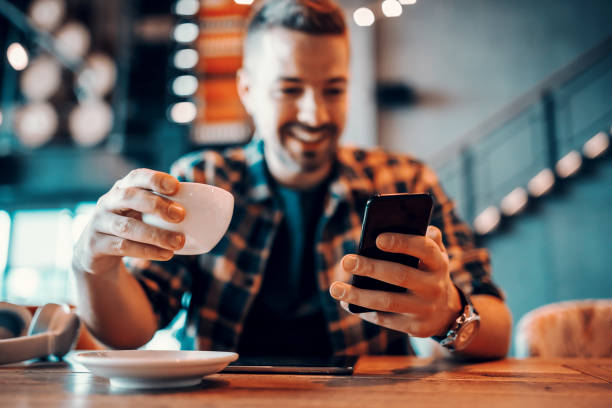 Smiling handsome Caucasian man in plaid shirt using smart phone and drinking coffee. Selective focus on hands, cafe interior.