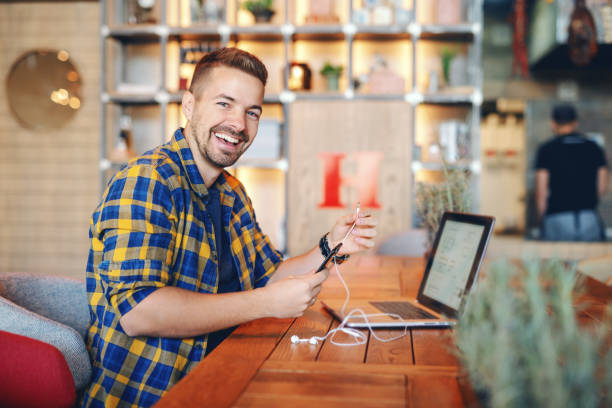 freelancer unshaven considerável caucasiano de sorriso na camisa de manta que põr auriculares no telefone esperto ao sentar-se no cafeteria. no portátil da mesa. - smart casual side view one person success - fotografias e filmes do acervo