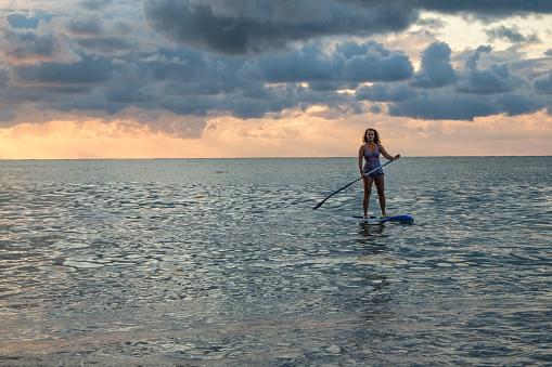 Young female paddling on SUP board in the sea at amazing dark sunset and waves.