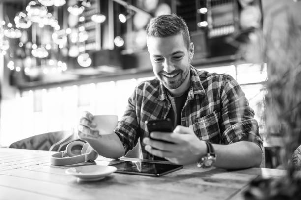Smiling handsome Caucasian man in plaid shirt using smart phone and drinking coffee. Selective focus on hands, cafe interior. Black and white photo.