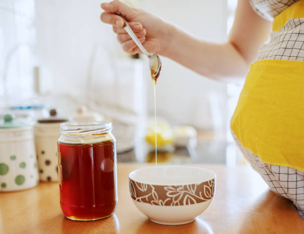 fermez-vous vers le haut de femme caucasienne enceinte dans le tablier mettant le miel du bocal dans le bol tout en restant dans la cuisine et préparant le petit déjeuner. - spoon honey cute jar photos et images de collection