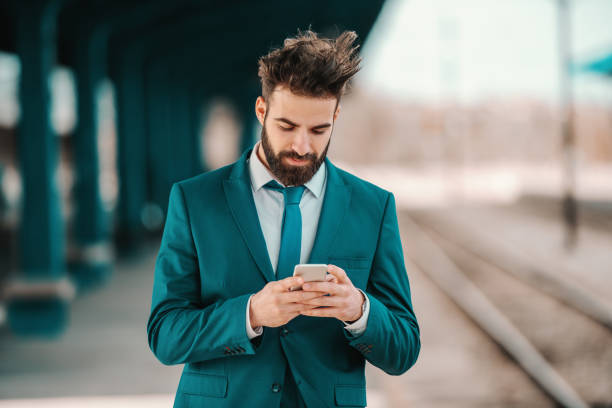 istock handsome Caucasian bearded businessman in turquoise suit using smart phone while waiting for train to arrive. Opportunities don't just happen, you create them. 1140818601