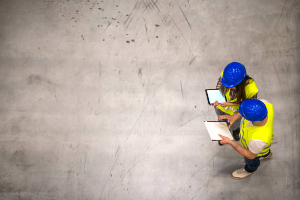 vue de dessus de deux ouvriers de construction portant des casques et des vestes réfléchissantes retenant la tablette et la liste de contrôle sur le fond gris de béton. groupe d’ingénieurs partageant des idées sur le projet. - construction checklist clipboard talking photos et images de collection