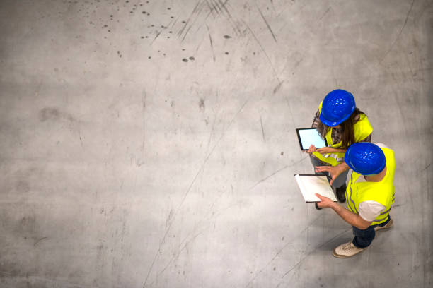 Top view of two construction workers wearing hardhats and reflective jackets holding tablet and checklist on gray concrete background. Group of engineers sharing ideas about the project. Group of engineers sharing ideas about the project. civil engineering photos stock pictures, royalty-free photos & images
