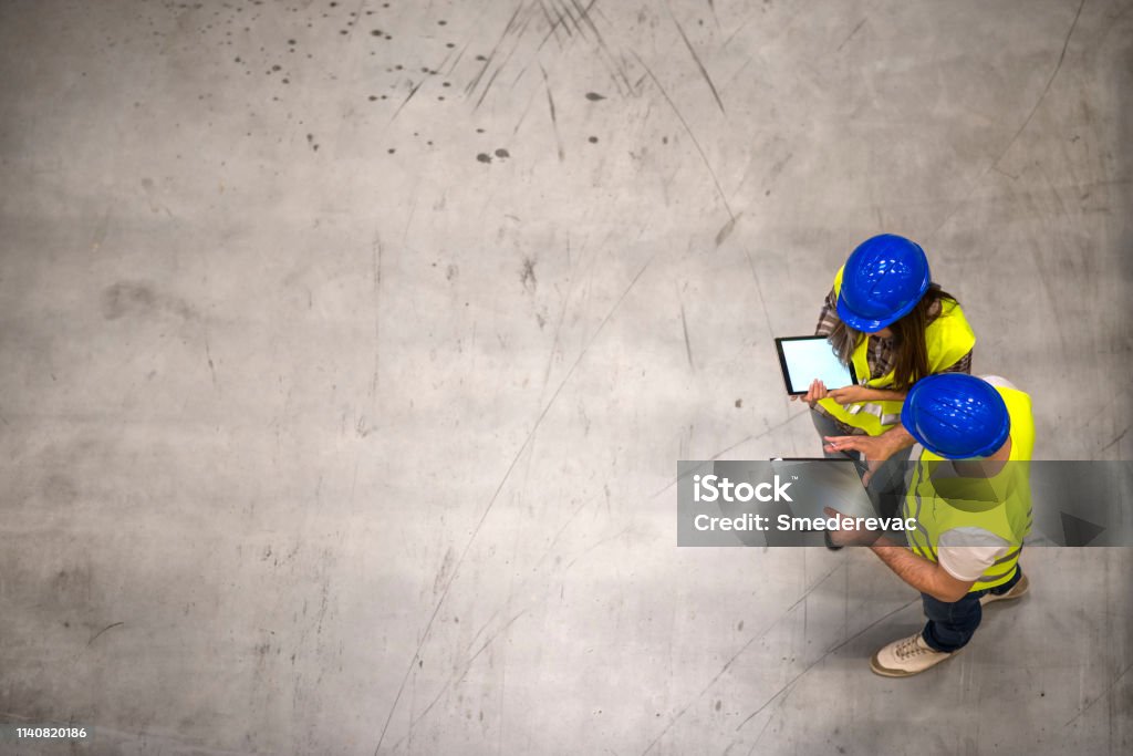 兩名穿著安全帽和反光夾克的建築工人在灰色混凝土背景上拿著平板電腦和清單的頂視圖。工程師小組分享有關該專案的想法。 - 免版稅建築地盤圖庫照片