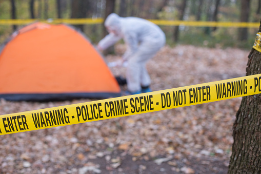 A scene of crime officer wearing a white boiler suit looks inside an examination tent at a crime scene. Crime scene in the forest