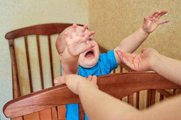 The little boy is crying in the crib and pulling his hands to his mom