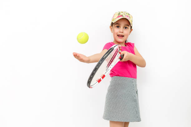 studio shot d’une petite fille souriante habillée en t-shirt rose et jupe blanche-sportswear pour le tennis sur fond blanc. elle se tient avec une raquette de tennis et jette une balle de tennis - tennis child teenager childhood photos et images de collection