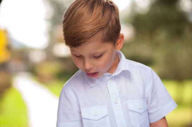 Portrait of a funny serious boy in a white shirt with a beautiful hairdo standing against the background of blurry trees outside the city.