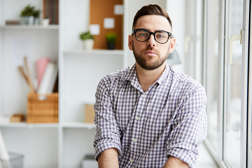 Bearded serious man in shirt and eyeglasses sitting by window in front of camera in office